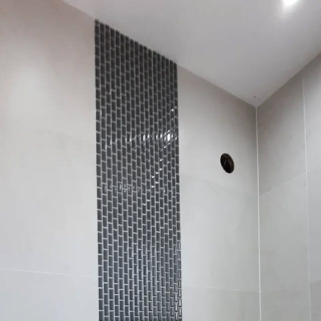 A white tiled bathroom wall with a narrow dark mosaic strip running from floor to ceiling, under a sloped ceiling.