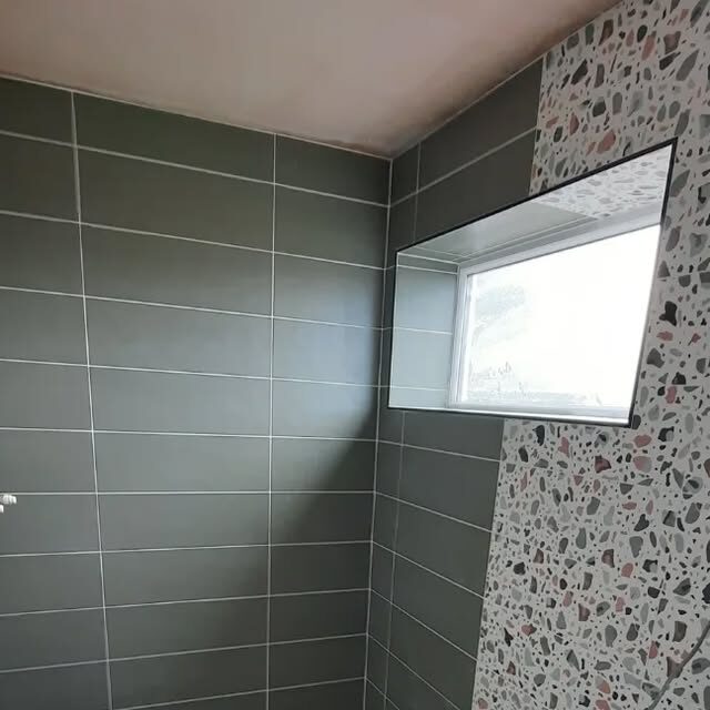 A bathroom tiled in sage green, with a terrazzo-effect panel running around the window reveal.