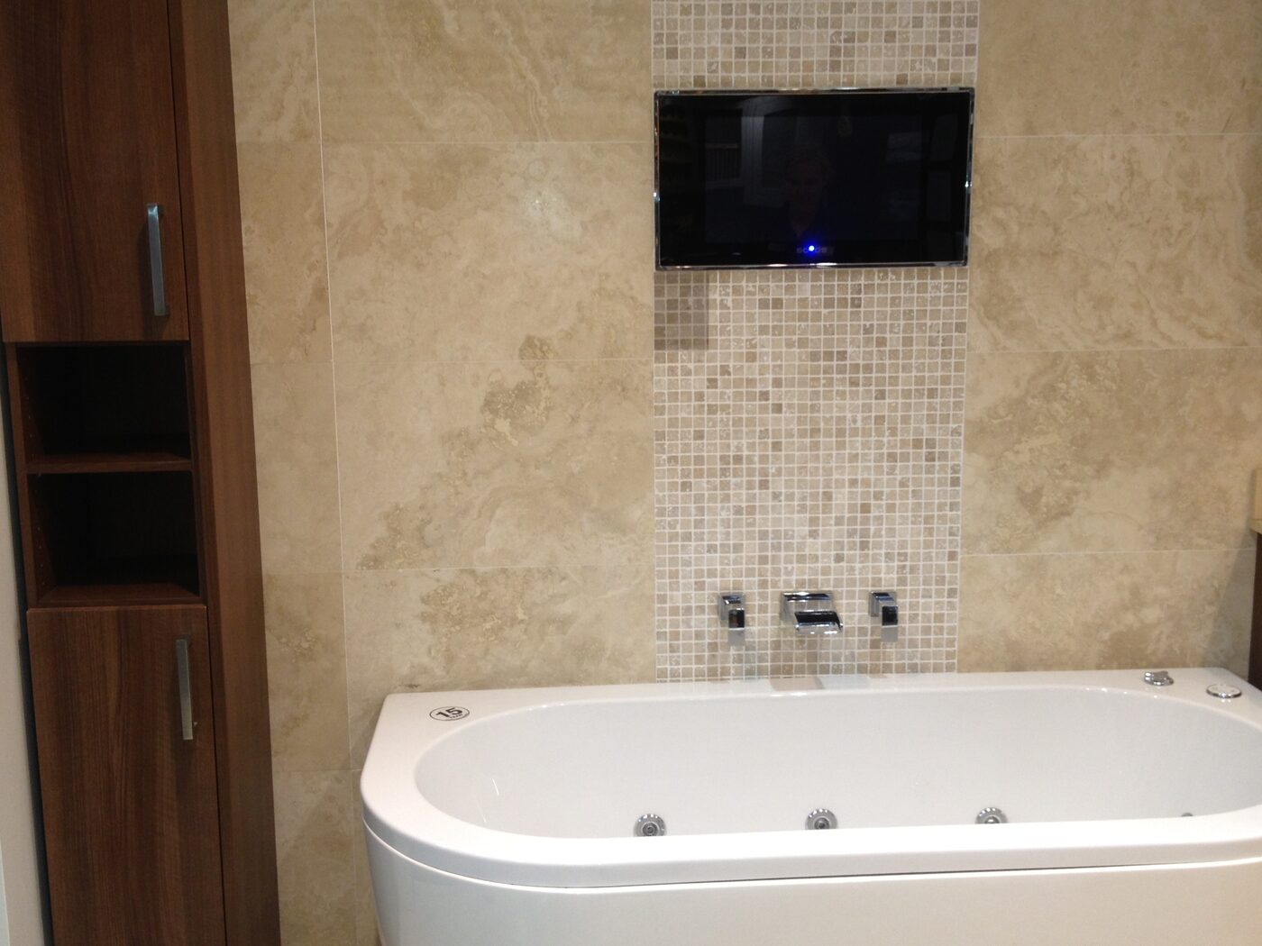 A curved white bath against a travertine-tiled wall, with a band of small mosaic tiles behind the taps and a television set into the tiling above.