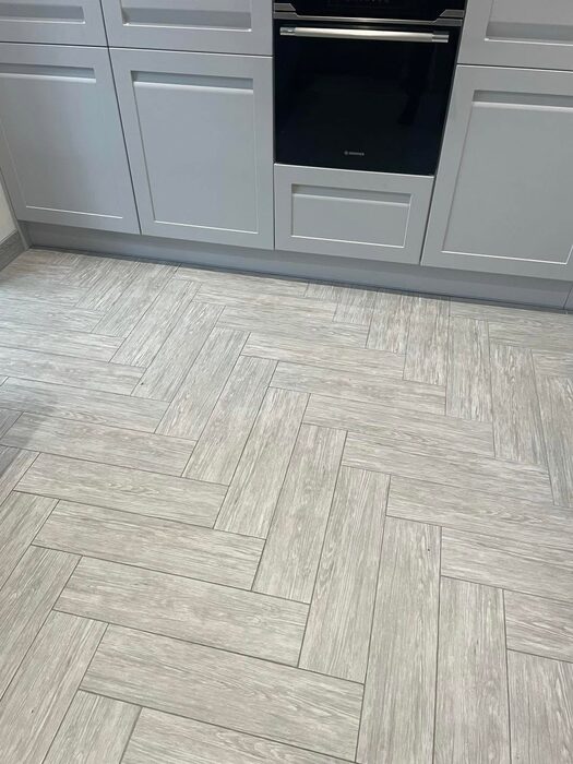 Pale wood-effect floor tiles laid in a herringbone pattern in front of grey kitchen units.