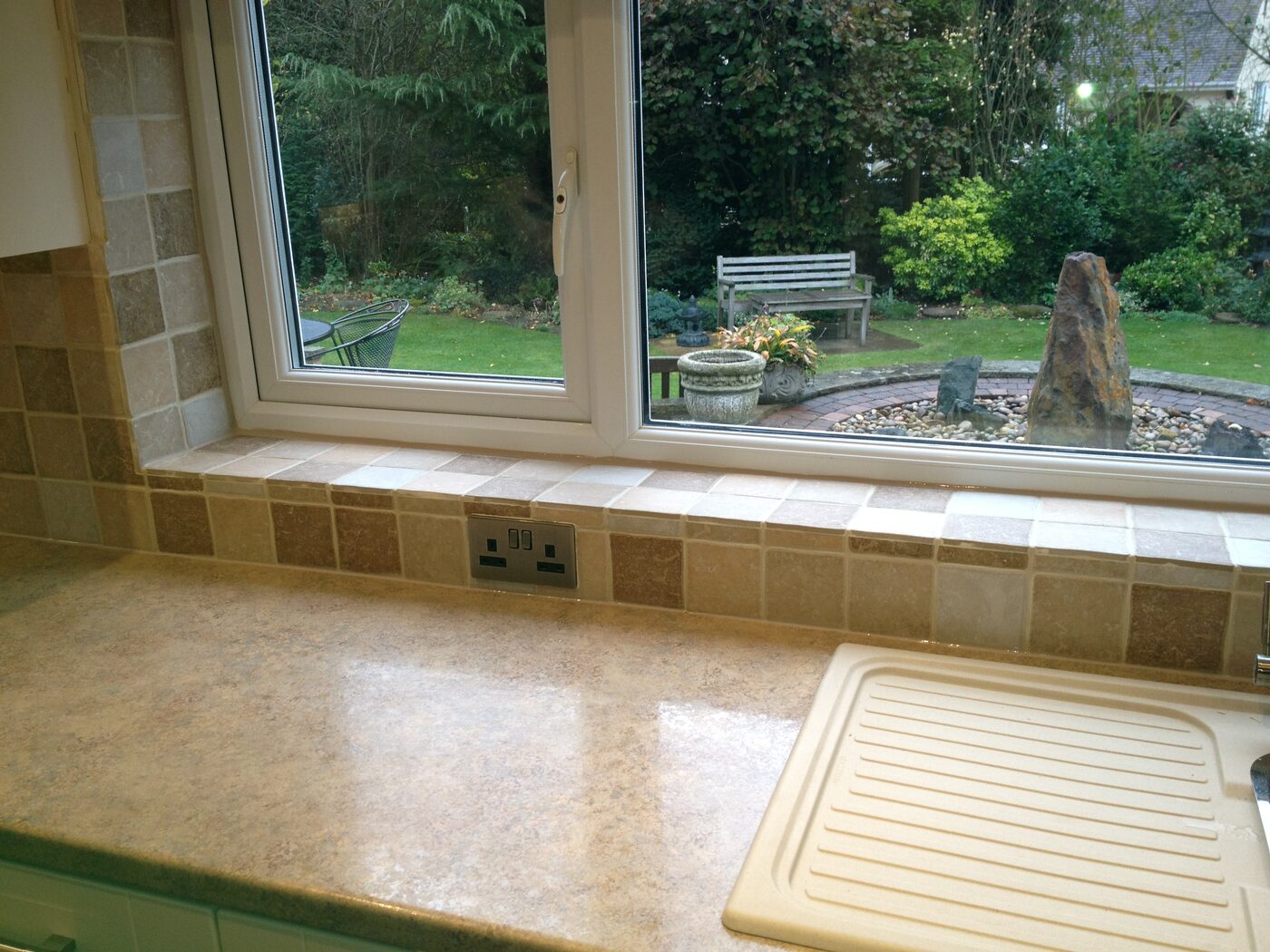 A kitchen window reveal tiled with a border of small square tiles in cream and brown, above a worktop, looking out onto a garden.