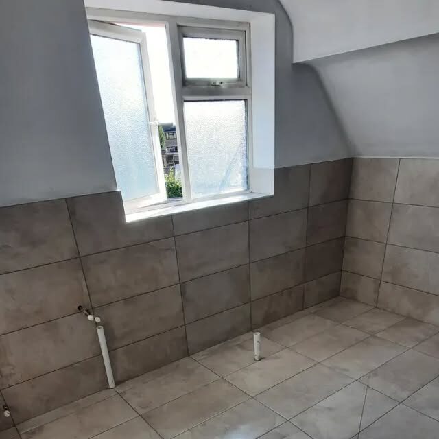 A loft bathroom part-tiled in grey tiles to half height under a sloped ceiling, with pipework still exposed at the wall.