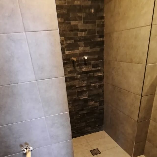 A shower enclosure tiled in large grey tiles with a dark bronze brick-effect feature wall at the back, and a drain set into the tiled floor.